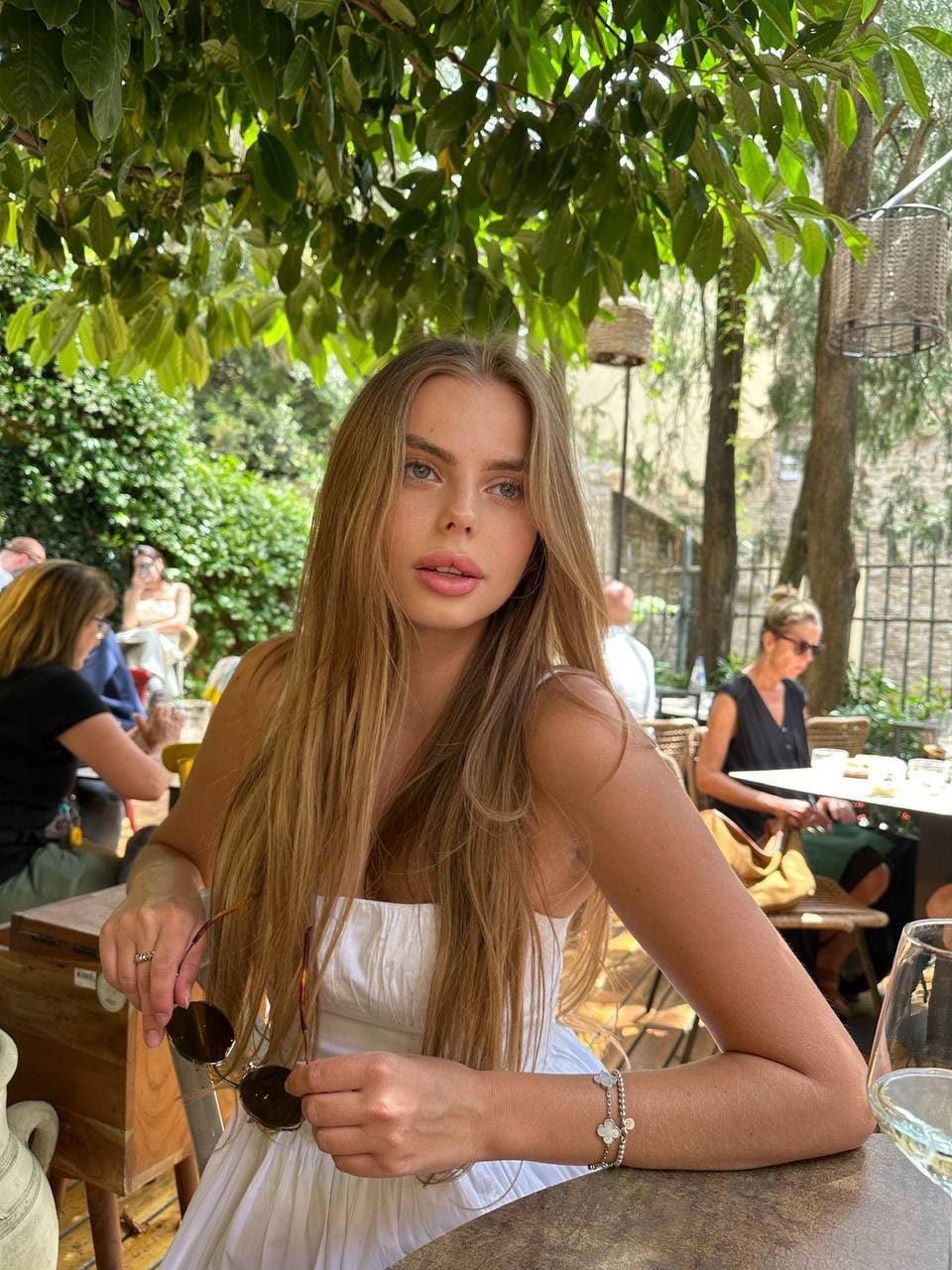 A young blonde woman sits at an outdoor table under trees, wearing a white strapless dress and holding sunglasses while looking toward the camera.