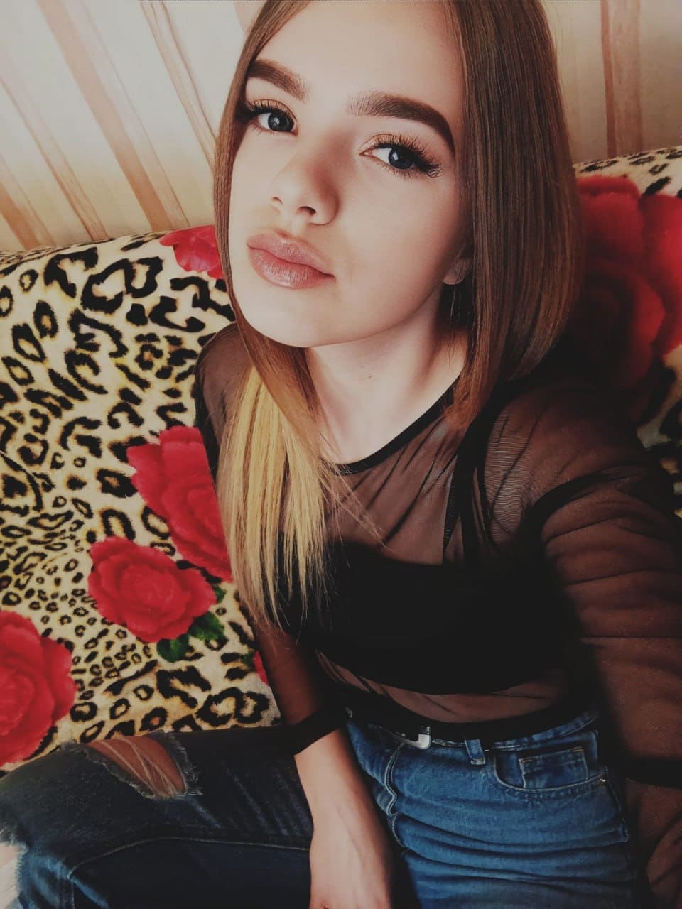 Mira sits on a leopard cushion with roses, wearing a sheer black top, bra, and ripped jeans.