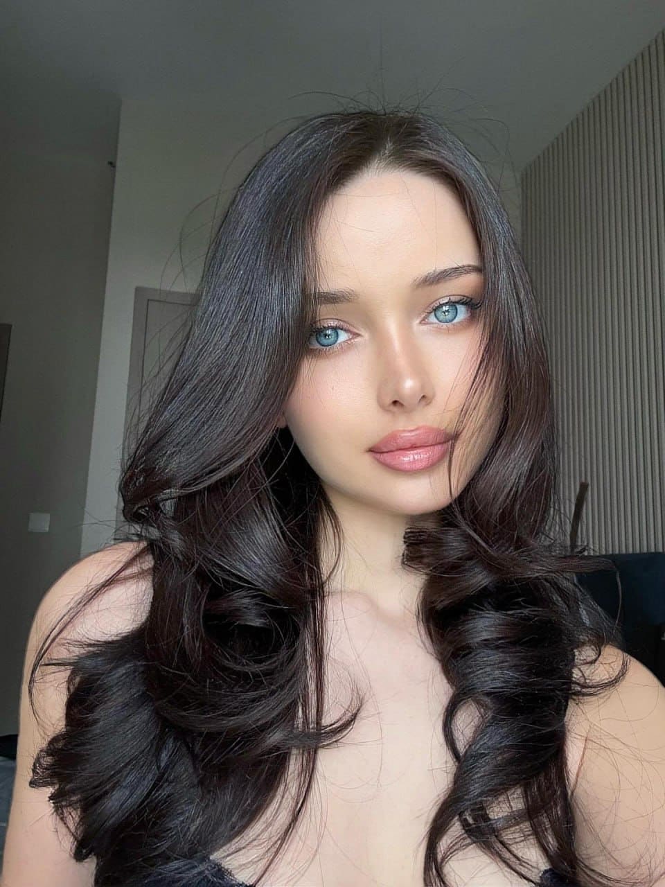 Portrait of a woman with wavy dark hair and blue eyes in a dark strapless top, set against an indoor background with a door and slatted panel.
