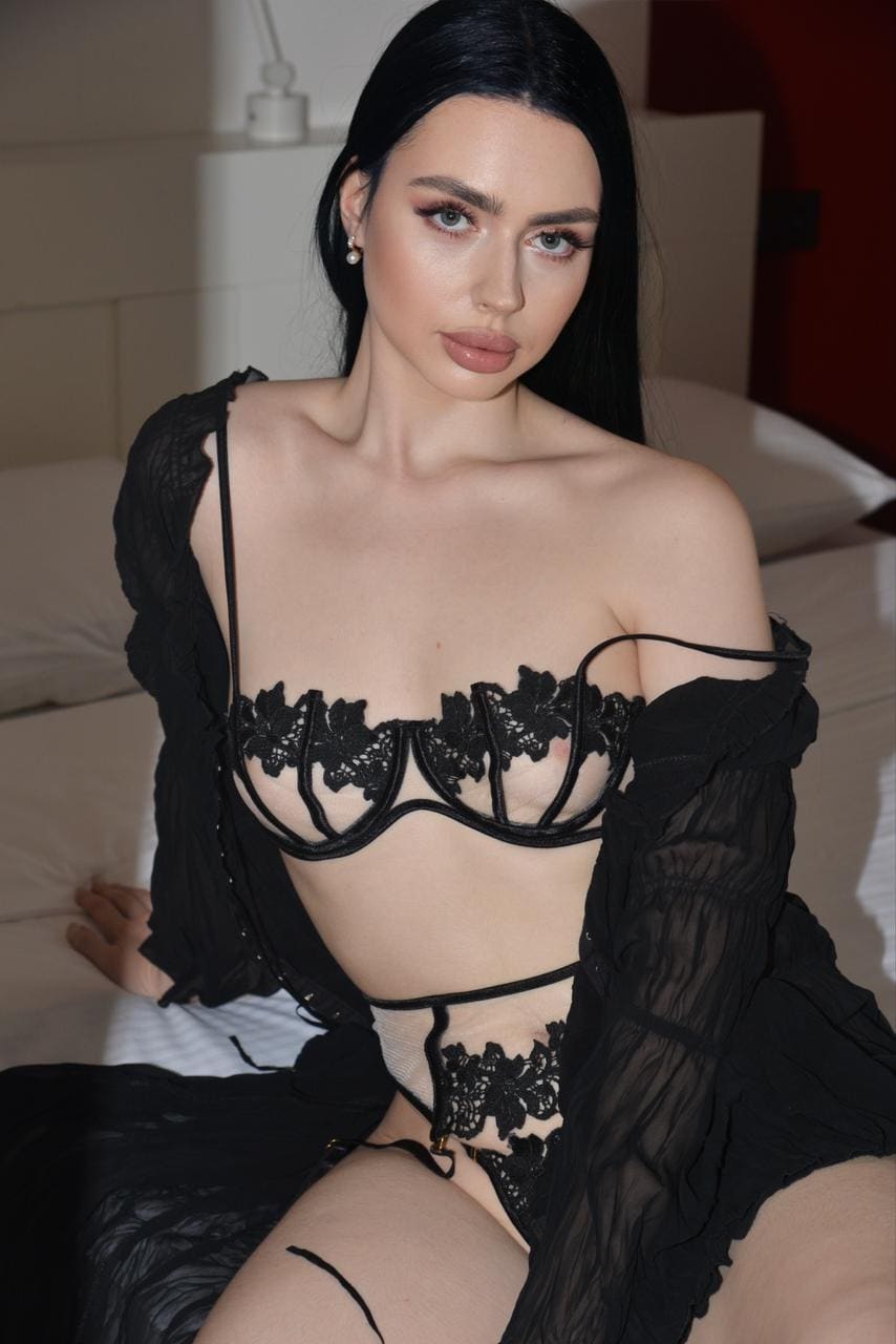 A woman sits on a bed in a black lace bra, garter belt, and sheer robe, with white bedding and a red wall behind her.