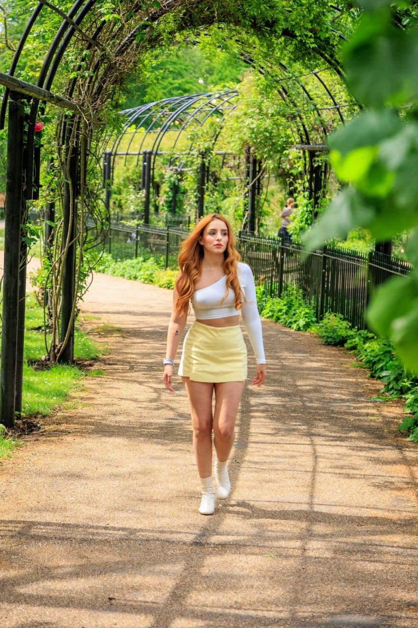 Walking on a gravel path under an arbor, she wears a white one-shoulder crop top, pale yellow skirt, and white ankle boots.