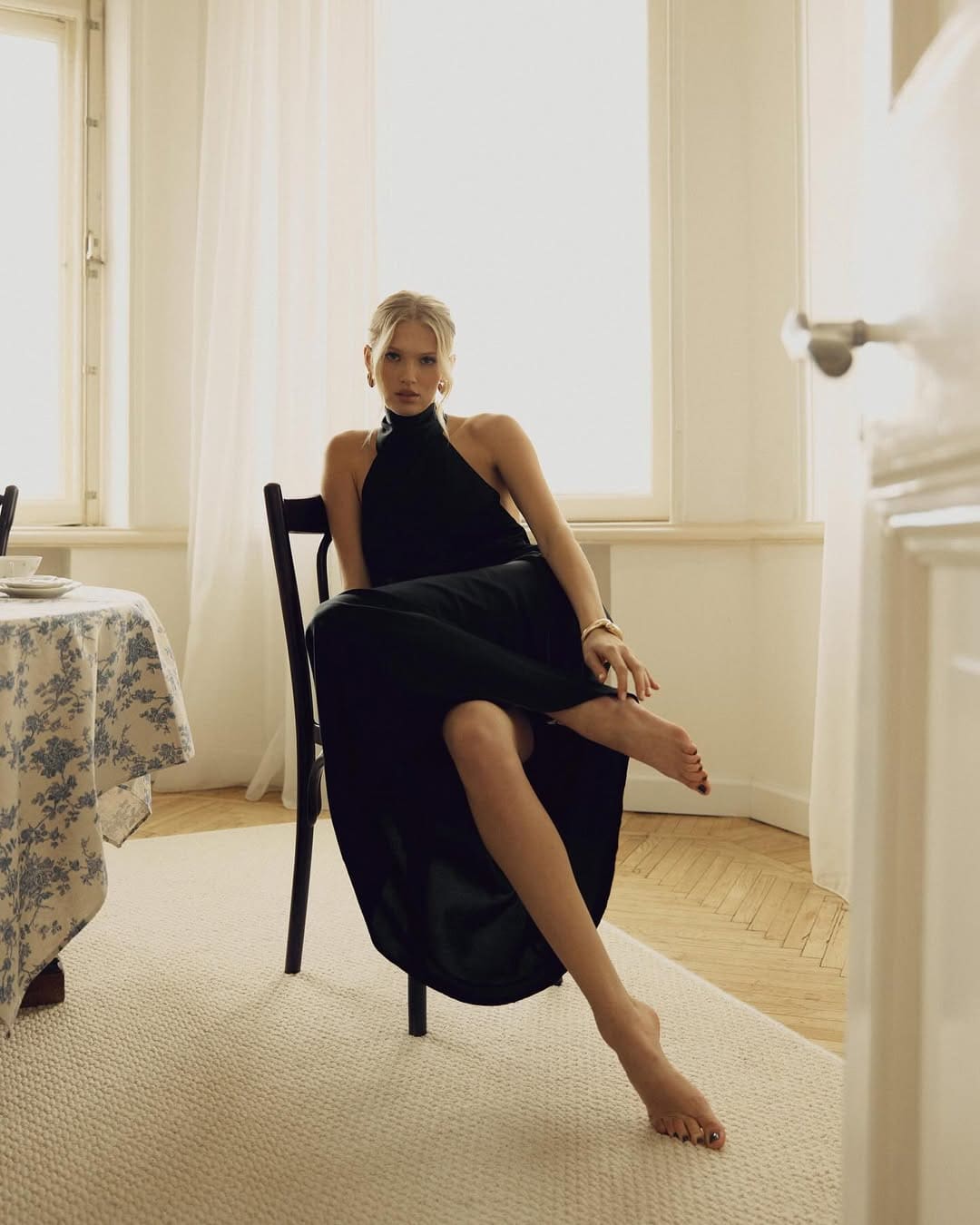 A blonde woman sits barefoot on a dark wooden chair in a long black halter dress, with a floral tablecloth and white curtains in the background.