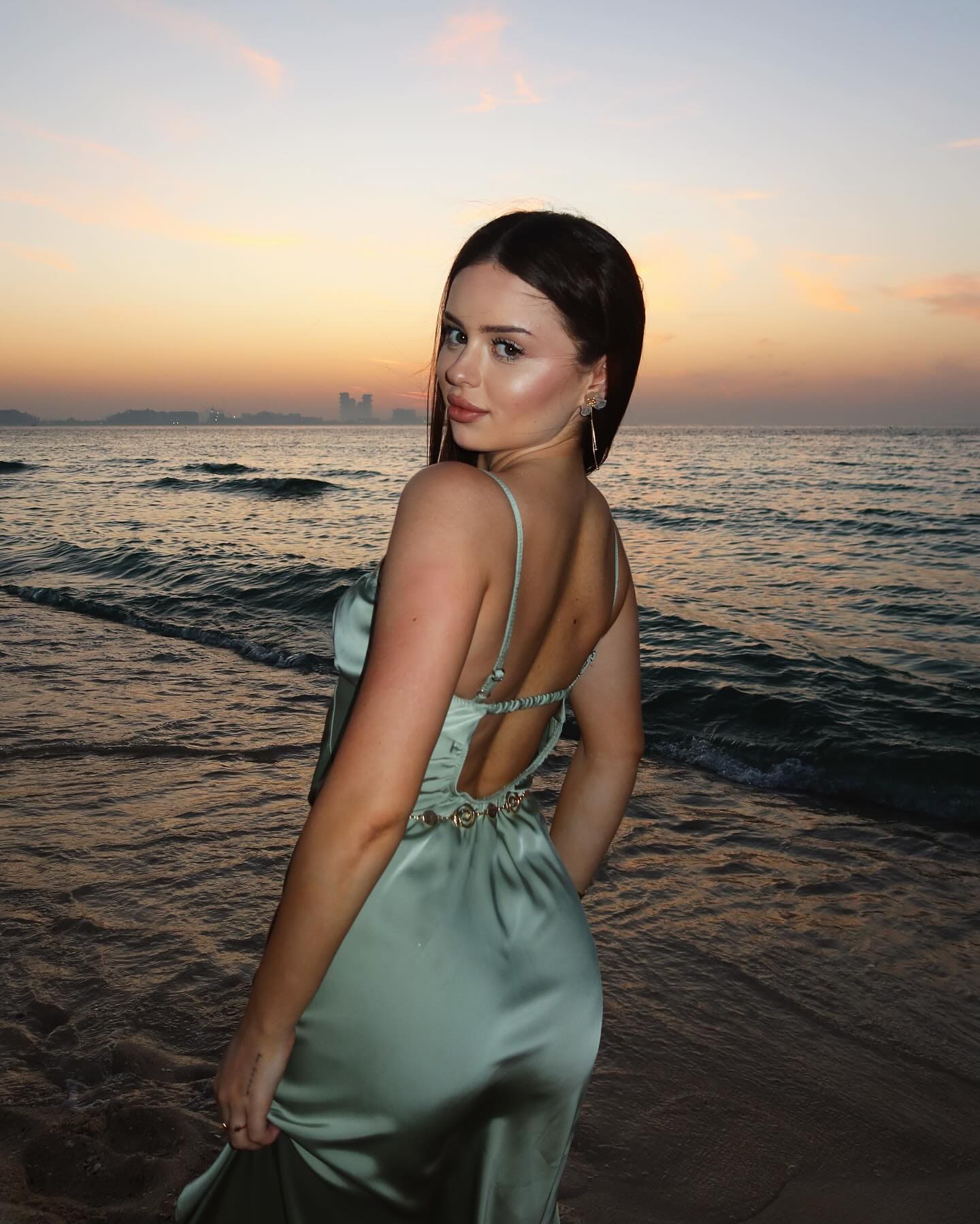 A woman in a light green satin dress with an open back stands on a beach at sunset, looking over her shoulder at the camera.
