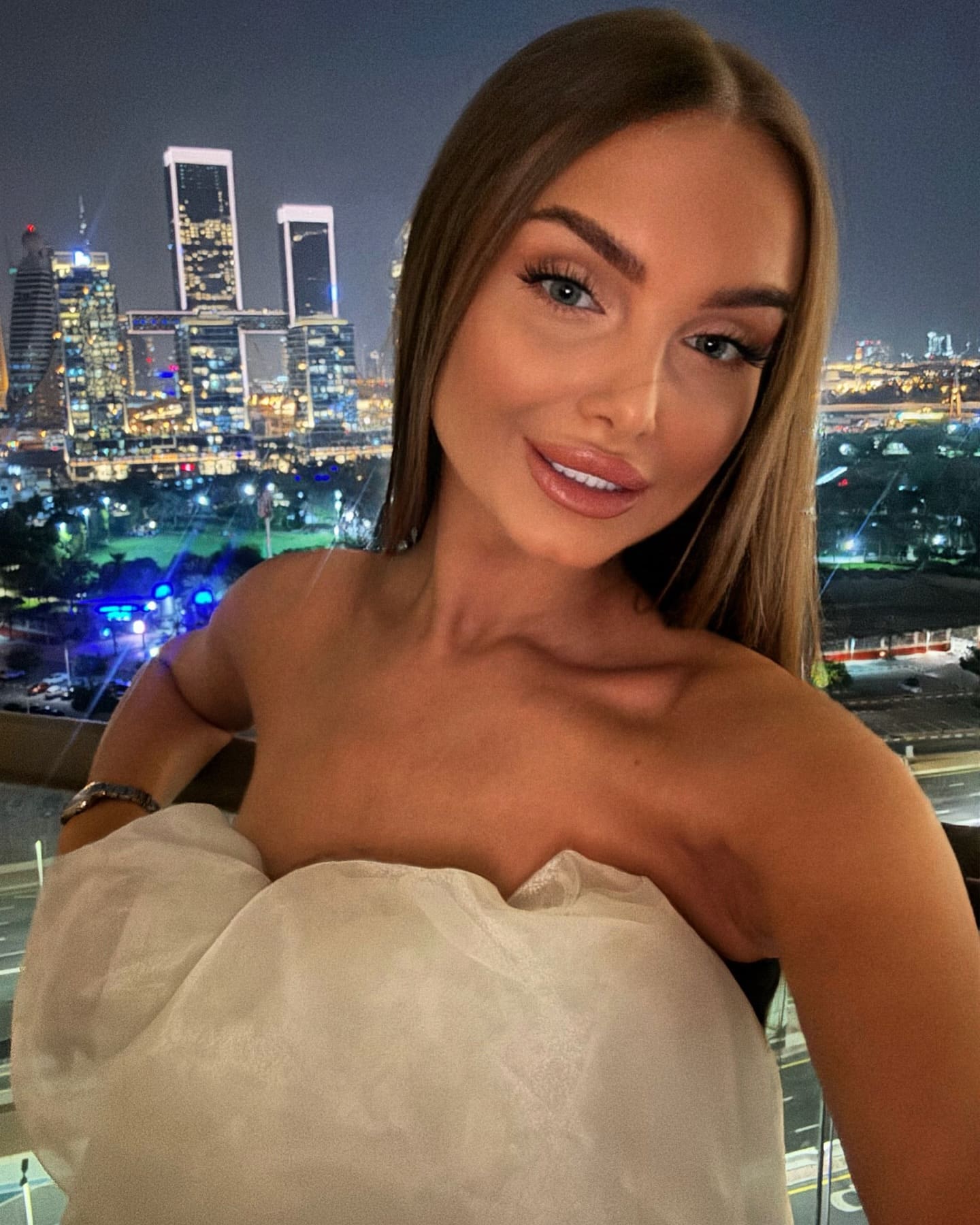 A woman in a white strapless top takes a selfie at night, with a lit city skyline visible in the background.