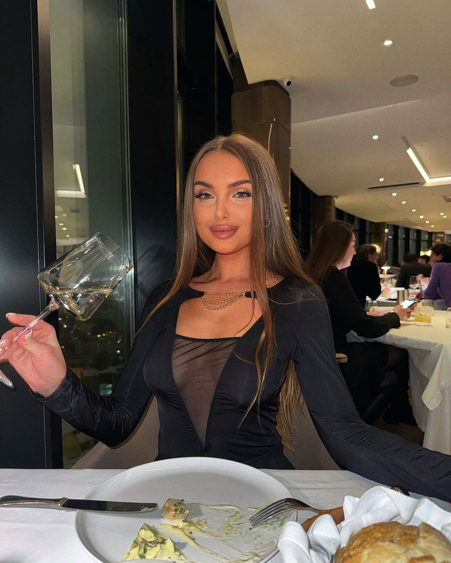 A woman in a black top with a sheer chest panel holds a wine glass at a restaurant table, with food and a bread roll on the white cloth.