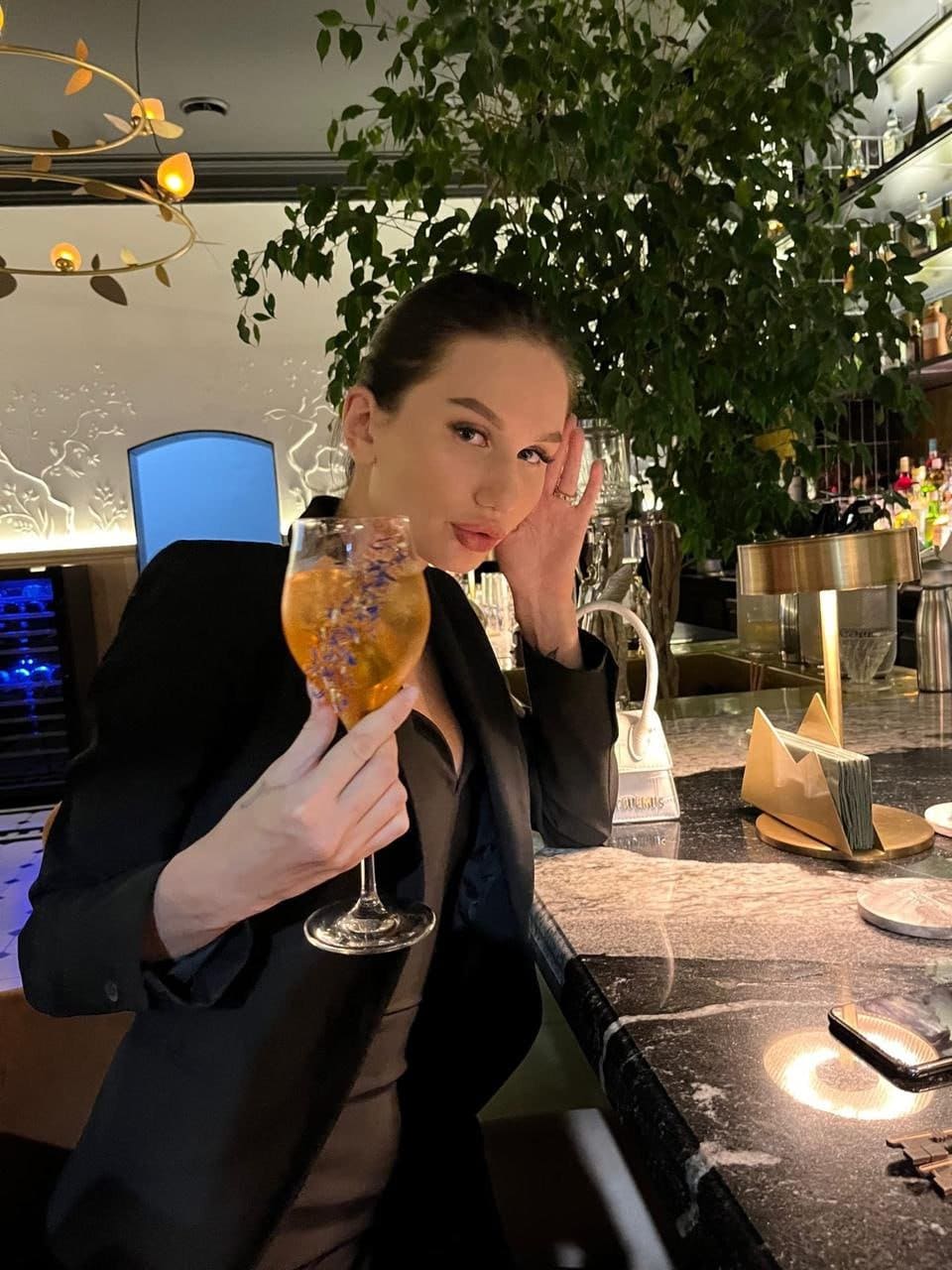 A woman in a black blazer sits at a marble bar, holding a stemmed glass with floral garnish in a dimly lit room.
