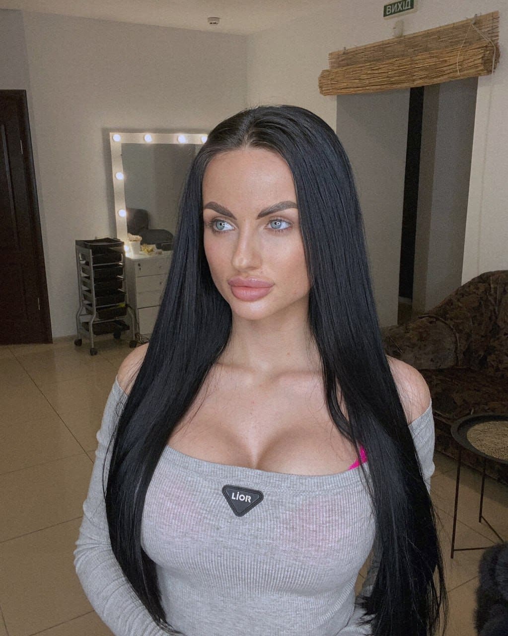 A woman stands in a room with a lighted mirror, wearing a grey off-the-shoulder ribbed top that reveals a pink strap underneath.