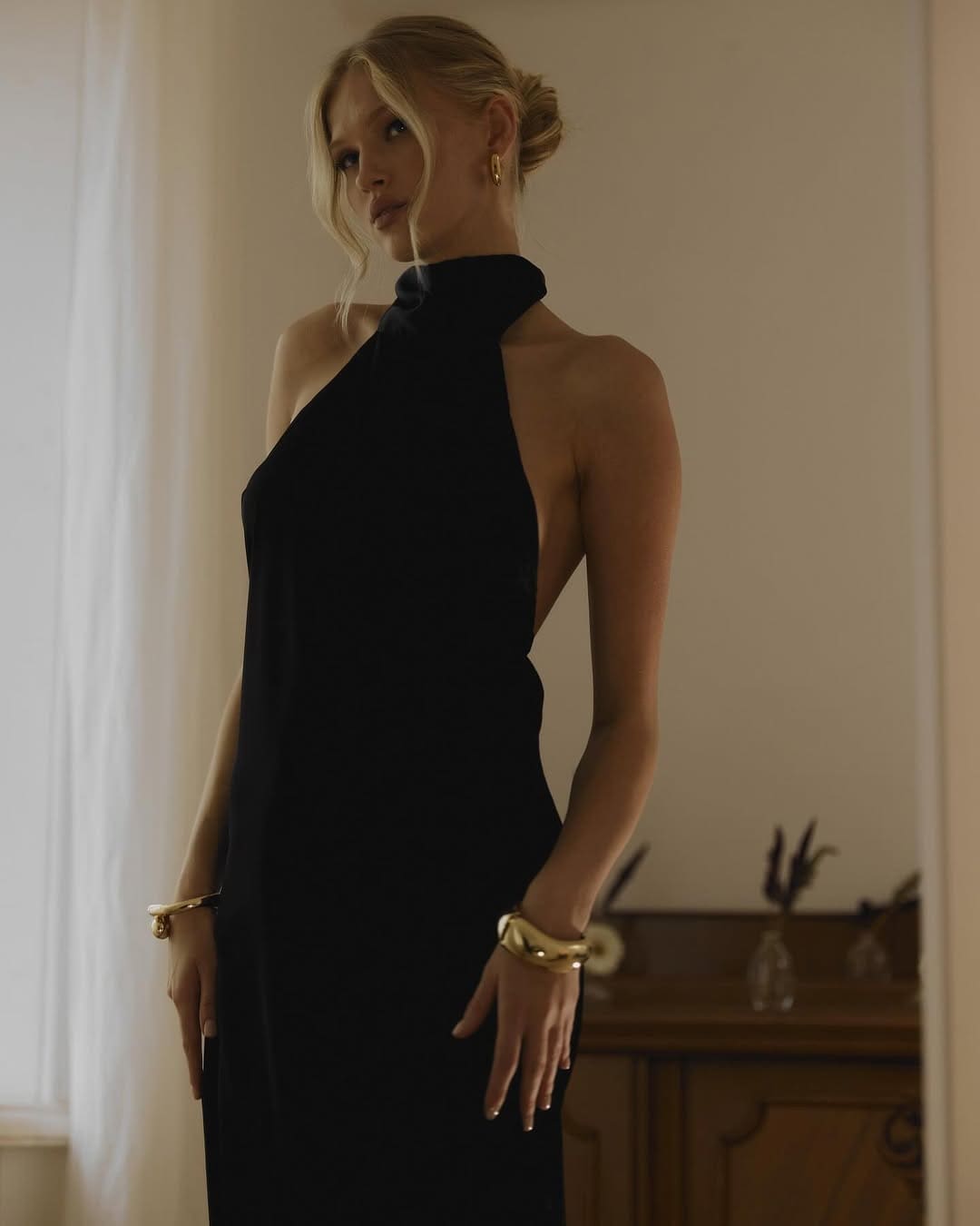 A blonde woman with a low bun stands in a black halter dress with a high collar and gold jewelry, in a room with a wooden cabinet and vases.