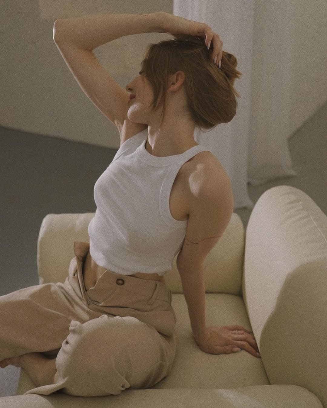 A woman with a loose bun sits on a cream sofa in a white cropped tank and beige trousers, one arm raised behind her head.