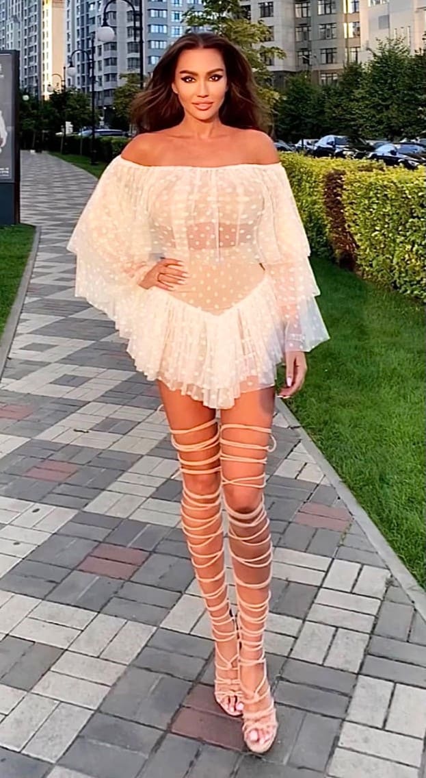 A woman stands on a paved walkway in a sheer off-the-shoulder polka-dot mini dress and lace-up thigh-high sandals, hand on hip.
