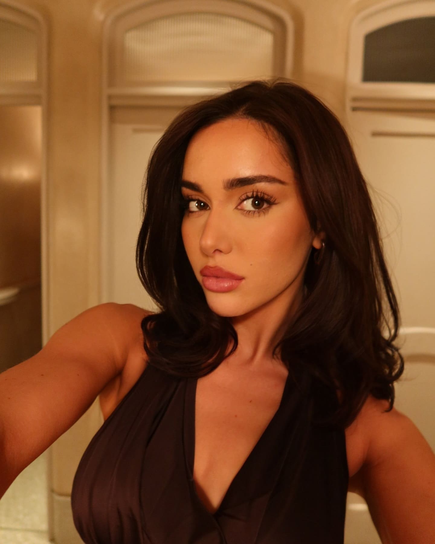 Elmaz stands indoors in a black sleeveless top with a deep V-neckline, posing before a paneled wall with arched details.