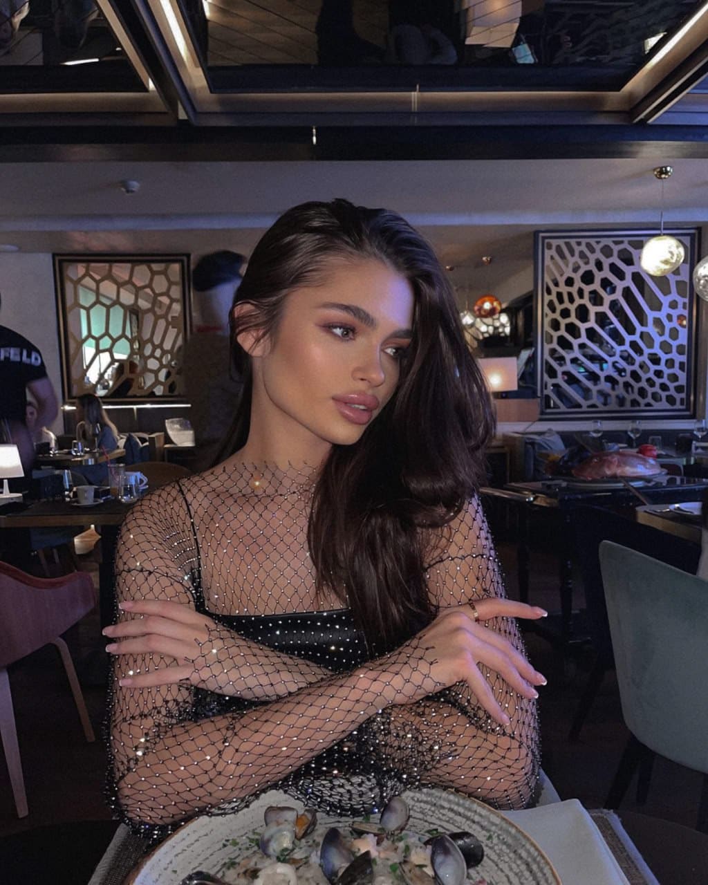 Woman seated at a restaurant table in a sheer beaded mesh top over black undergarment, arms crossed, with a plate of mussels.
