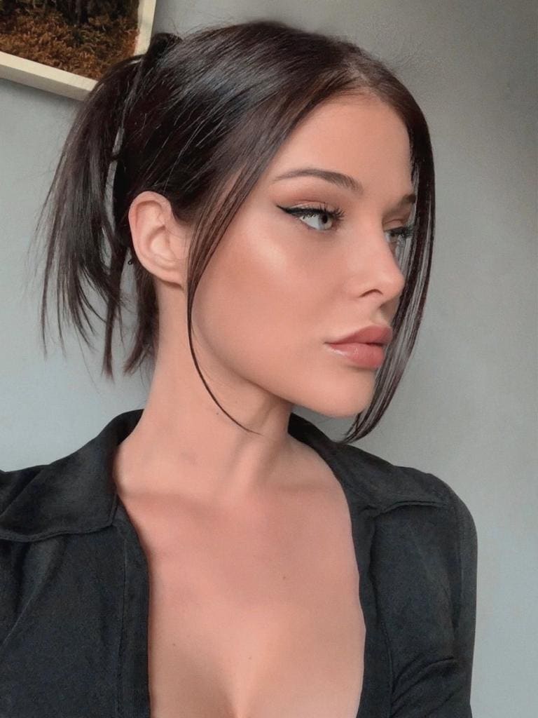 A woman with a high ponytail wears a black collared shirt with an open neckline against a gray wall with dried plants.