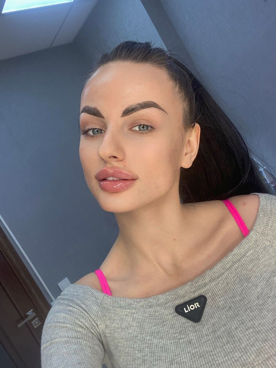 A close-up selfie of a woman with a ponytail in a grey off-shoulder top with pink straps, against a grey wall and wooden door.