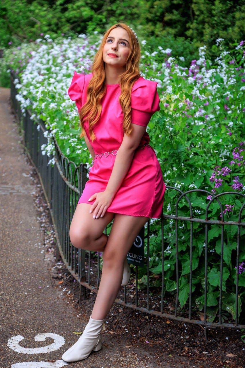 On a paved path by a black fence, she wears a short pink dress with puffed sleeves and a heart belt, one leg crossed, head tilted up.