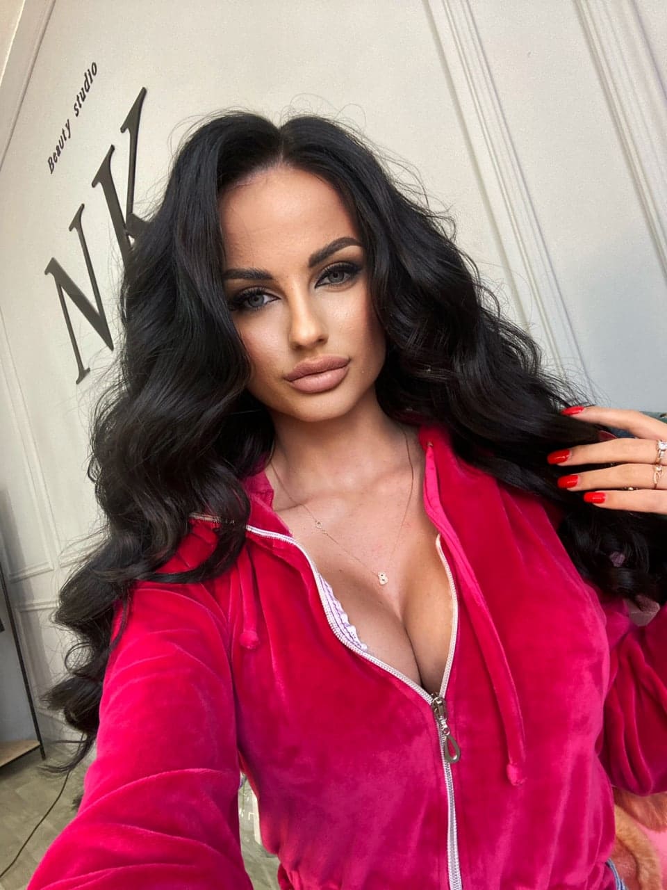 A selfie shows a woman in a pink hoodie, unzipped to reveal a white lace bra, with a hand near her hair and large letters on the wall.