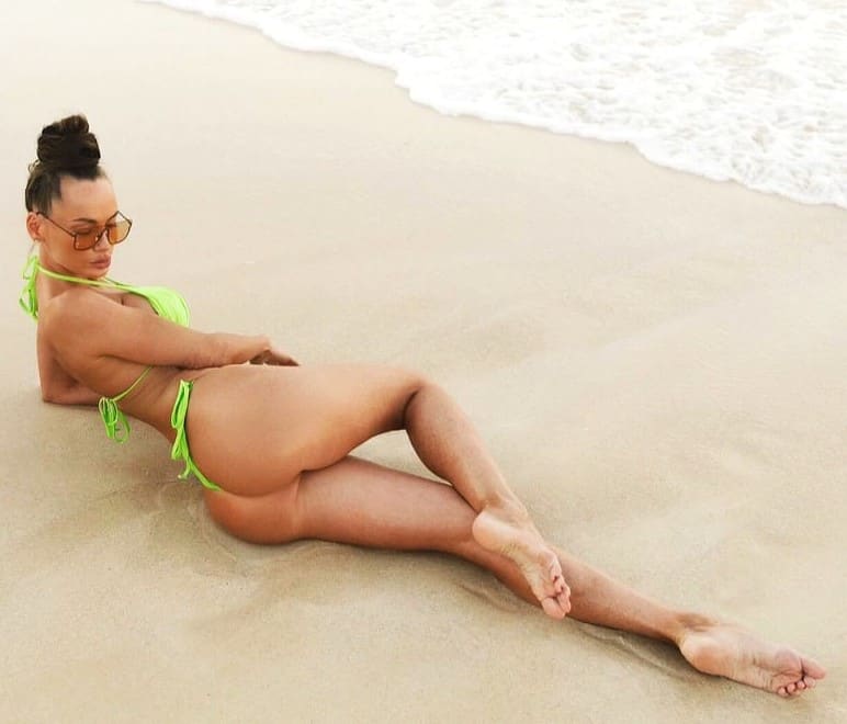 A woman in a bright green string bikini lies on her side on sandy beach, wearing sunglasses, with ocean waves in the background.