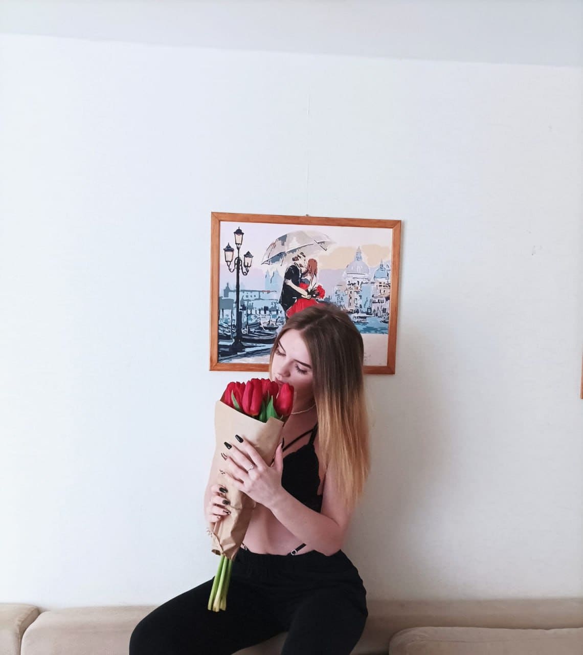 A woman in a black crop top holds red tulips to her face on a beige couch, with a painting behind.