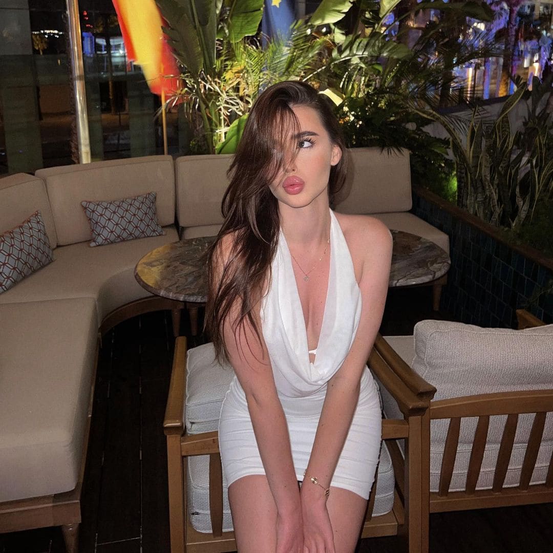 Seated on a wooden chair at night, a woman wears a white halter mini dress with a deep V-neck, looking to the side.