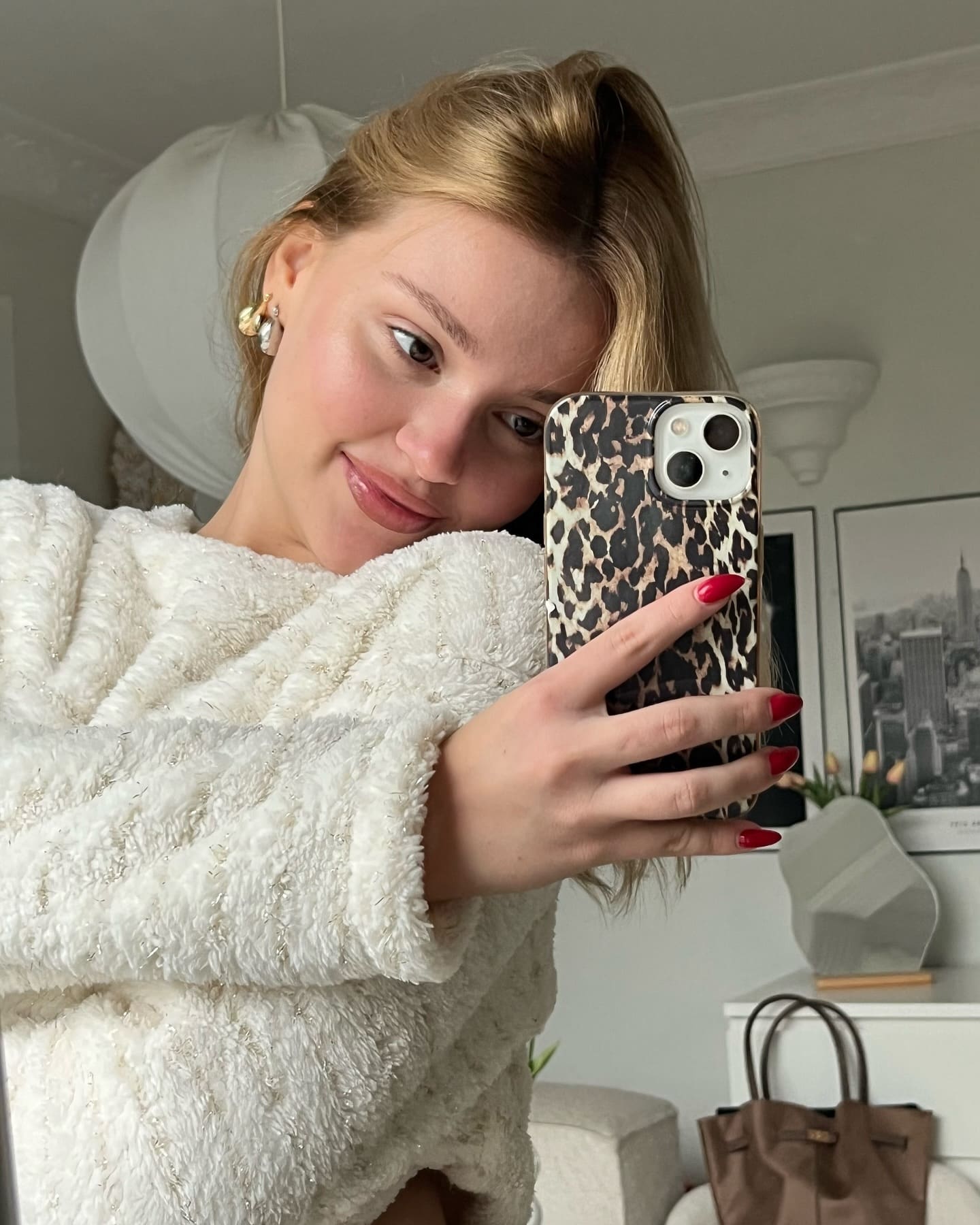 A blonde woman with a high ponytail takes a mirror selfie in a white fuzzy sweater, holding a phone with a leopard-print case.
