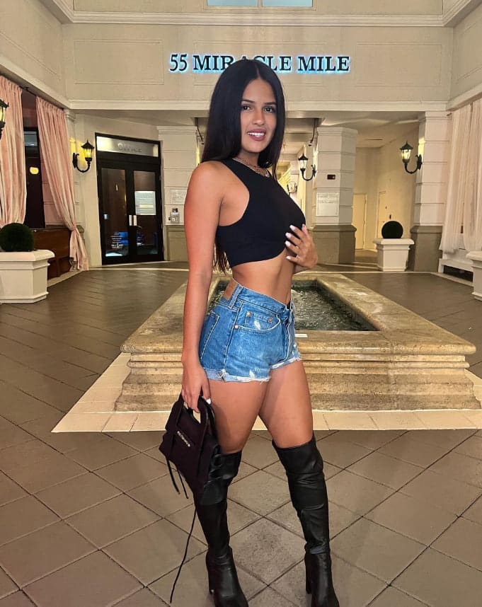 A woman stands in a tiled lobby with a fountain and '55 MIRACLE MILE' sign, wearing a black crop top, denim shorts, and knee-high boots.