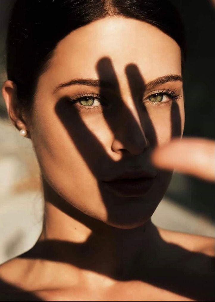 A woman with dark hair pulled back holds a hand before her face, casting finger shadows on her eyes and nose.
