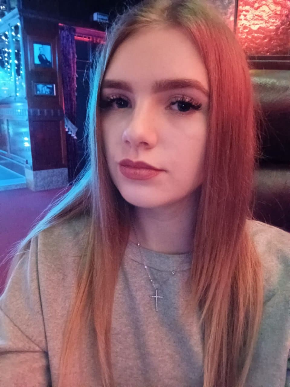 A woman with a cross necklace sits indoors in a grey top, against a background of blue and red lighting.
