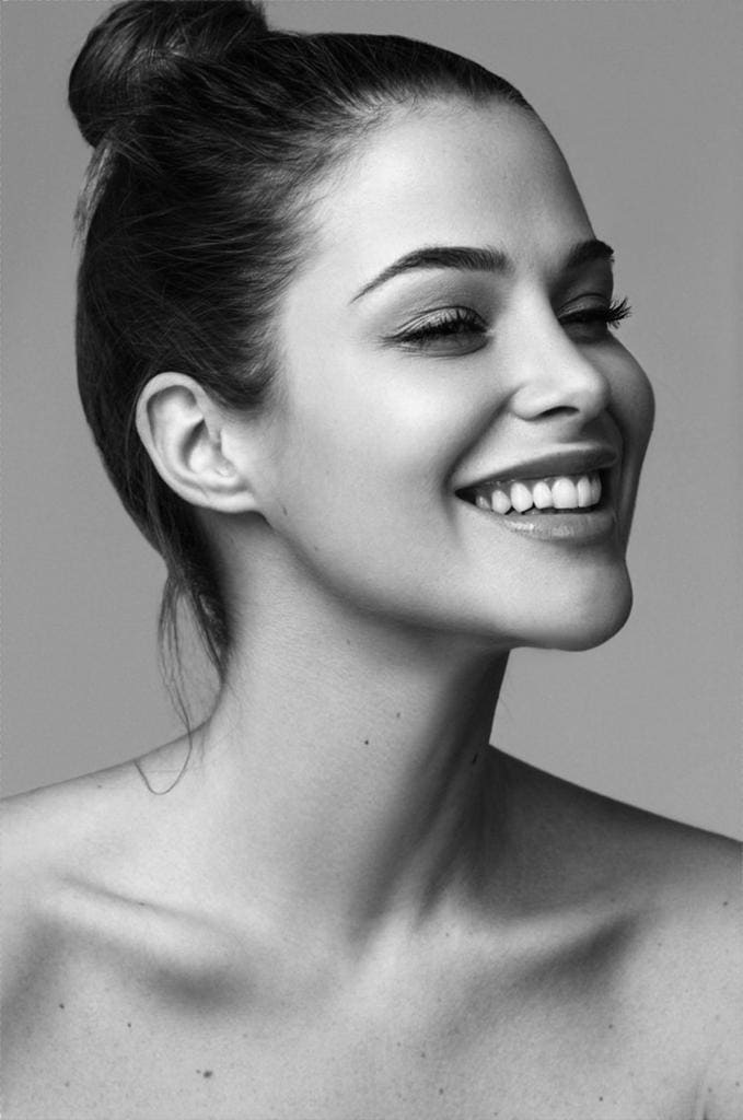 A black-and-white portrait shows a woman with a high bun smiling, bare shoulders visible against a gray background.