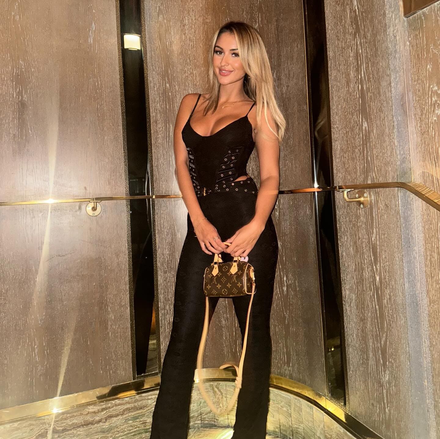 Arsenia stands in a wood-paneled elevator wearing a black sleeveless top and pants, holding a small patterned handbag.