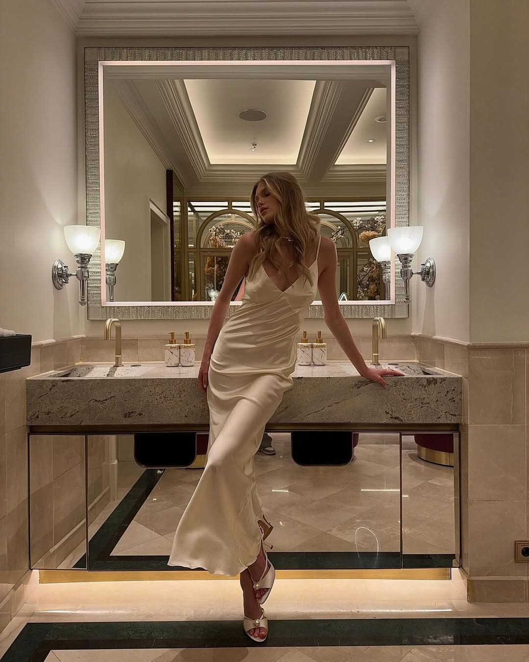 A blonde woman in a floor-length cream satin dress and white strappy heels leans against a marble counter in a bathroom with a large mirror and sconces.