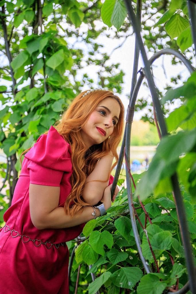 Leaning against a leafy trellis, she wears a magenta dress with a heart belt and pearl headband, looking toward the camera in a garden.