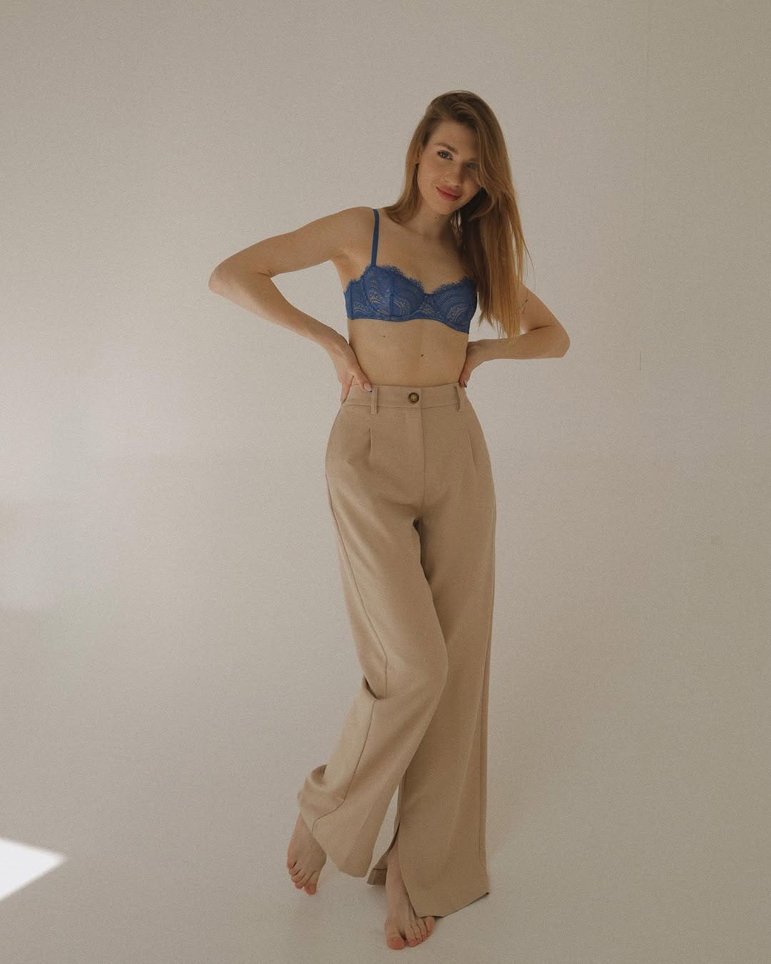 A barefoot woman with light-brown hair wears a blue lace bra and beige wide-leg trousers, hands on hips, one leg crossed against a light wall.