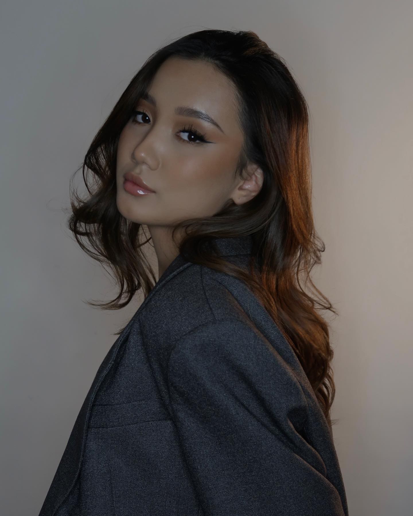 Timina, with long wavy dark hair, wears a grey blazer and looks over her shoulder against a plain light wall.