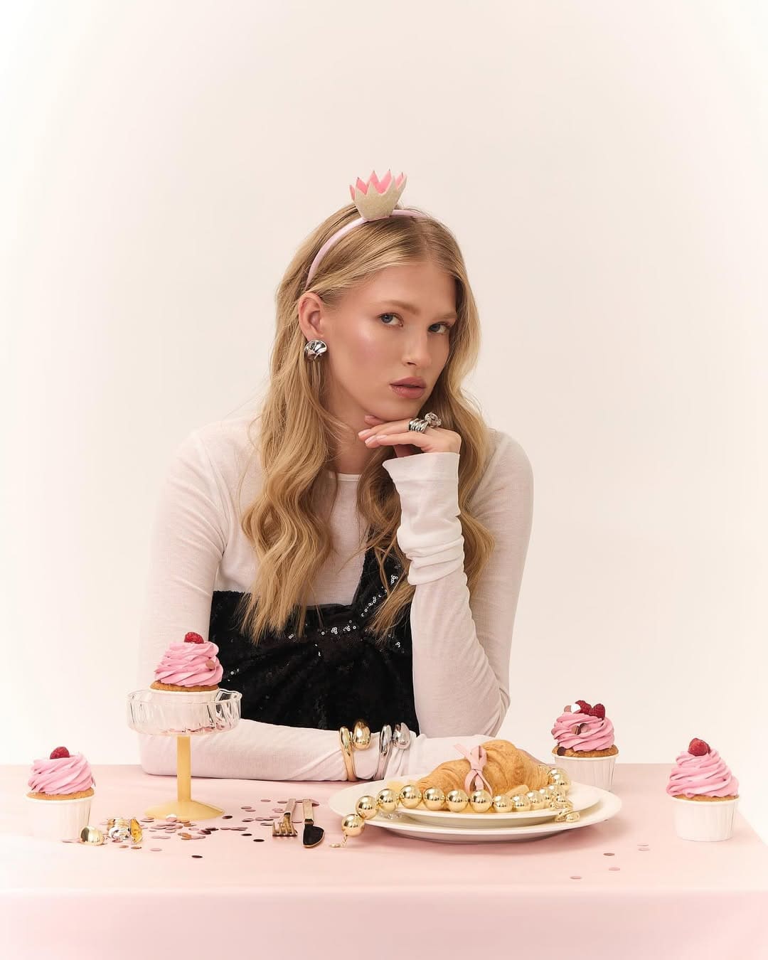 Caroline sits at a pink table in a black sequined dress and white top, with a crown headband, rings, and cupcakes on a plate before her.